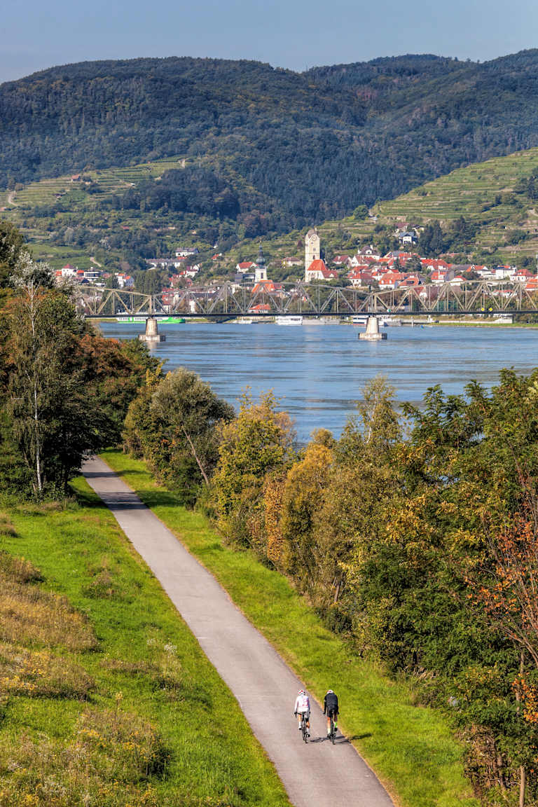 A scenic landscape with a winding path in the foreground, surrounded by lush greenery and a picturesque town nestled along the shores of a tranquil body of water, with mountains rising in the background.