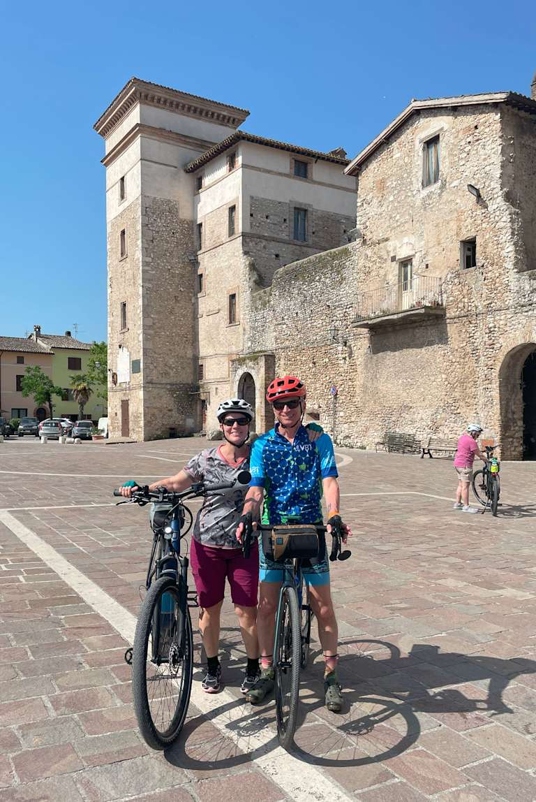 The image shows two cyclists posing in front of a historic stone building with a square tower, surrounded by a paved plaza and other buildings in the background.