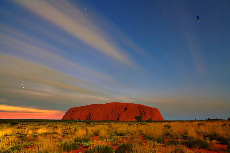 A majestic, red-hued rock formation stands prominently against a vibrant sky filled with colorful streaks of light, set amidst a vast, arid landscape.