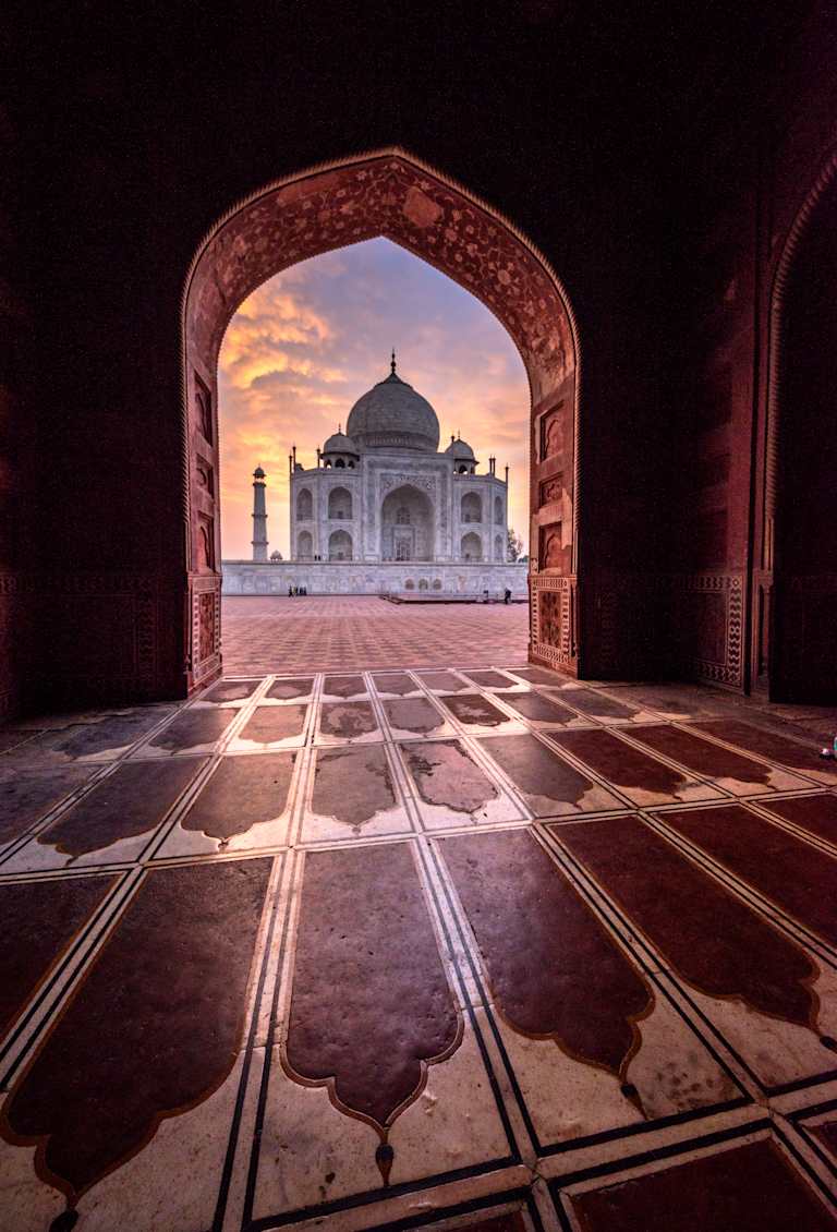 A magnificent domed structure, the Taj Mahal, is framed by an ornate archway, with the reflection of the building visible on the intricate tiled floor in the foreground.