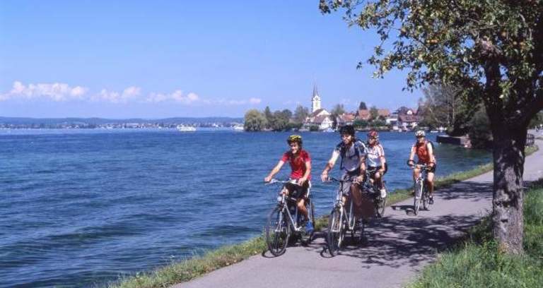 Three cyclists riding along a path next to a scenic lake, with a town and church steeple visible in the background.