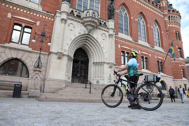 A person on a bicycle rides in front of a grand, ornate brick building with arched windows and intricate architectural details.