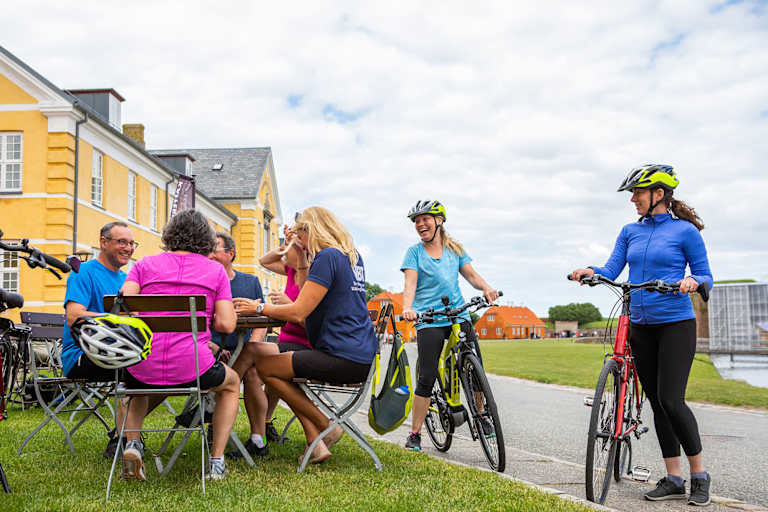 A group of people, some on bicycles and others seated, gathered in an outdoor setting with colorful buildings in the background.