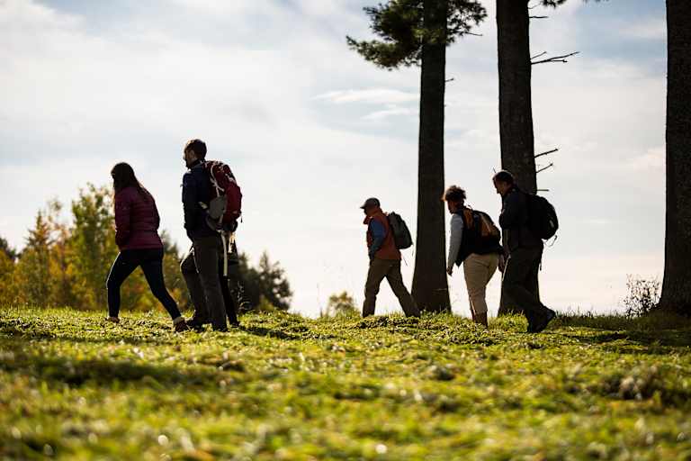 A group of people standing on a grassy hill, with tall trees in the background against a cloudy sky.