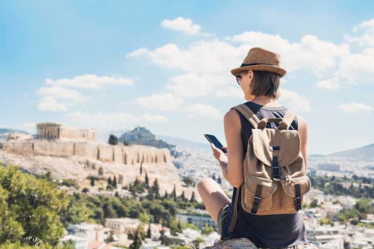 A person wearing a hat and backpack stands facing a scenic view of ancient ruins and buildings against a backdrop of cloudy blue sky.