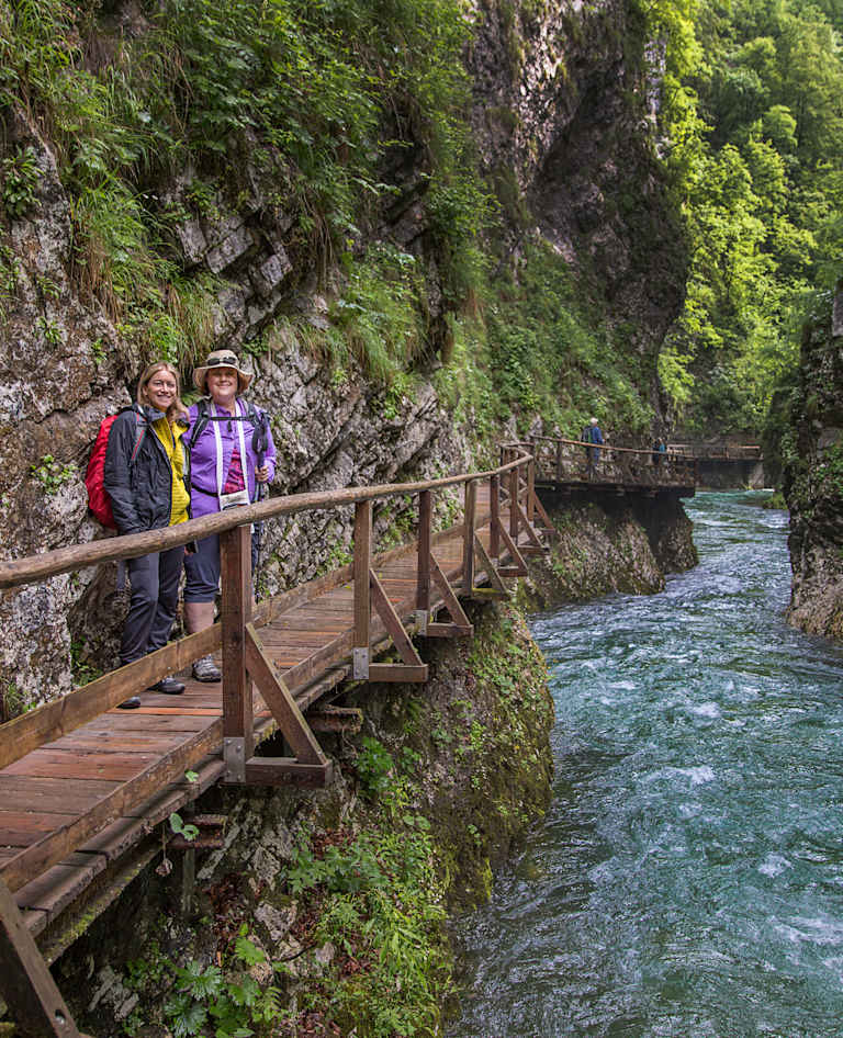 A wooden walkway winds along a rocky, moss-covered cliff face, with a rushing river flowing below, surrounded by lush, verdant foliage.