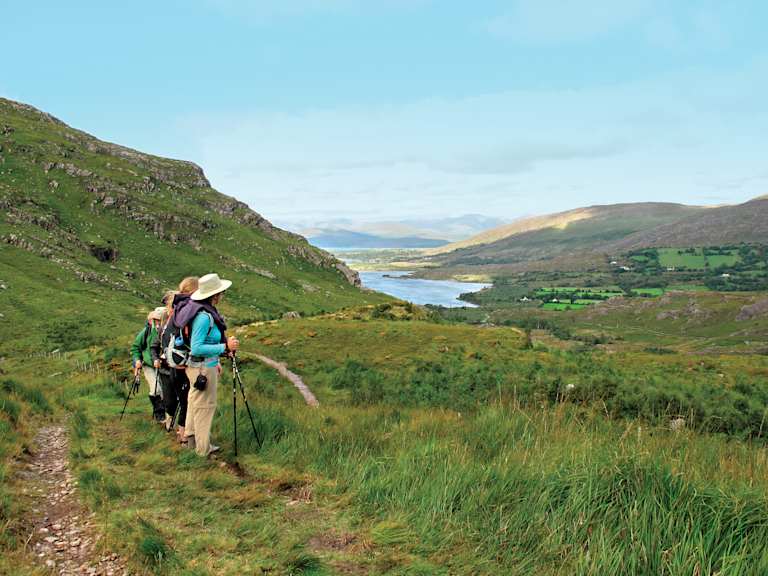 A hiker walks along a grassy trail in a scenic mountainous landscape, with a lake and distant hills visible in the background.