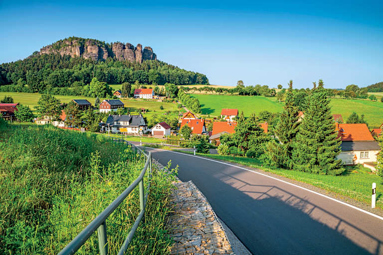 A picturesque village nestled in a lush, green valley with towering rock formations in the background, connected by a winding road.