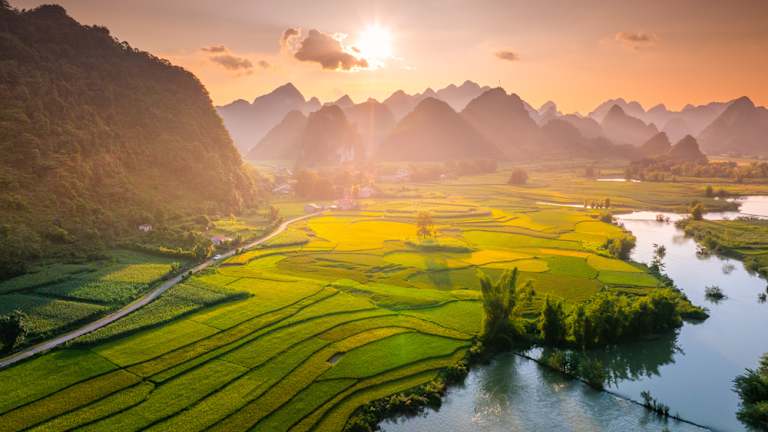 Lush green rice terraces stretch out across the landscape, framed by towering mountains and a glowing sunset sky.