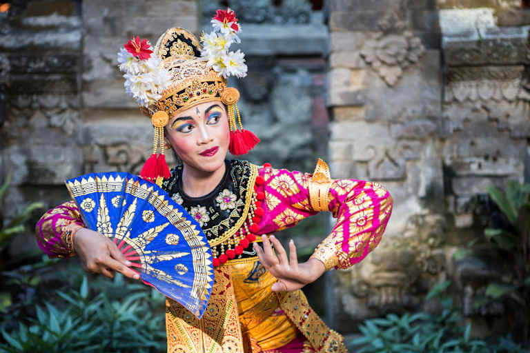 A colorfully dressed performer with an elaborate headdress and fan poses in front of a stone wall backdrop, surrounded by lush greenery.