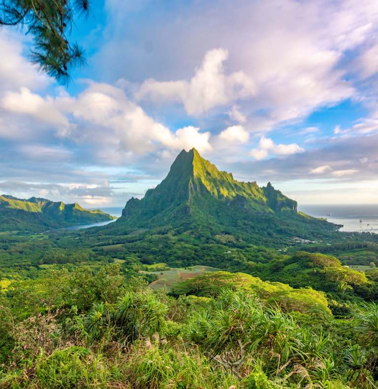 A lush, verdant landscape with a towering, rugged mountain peak rising in the distance, surrounded by a dramatic cloudy sky.