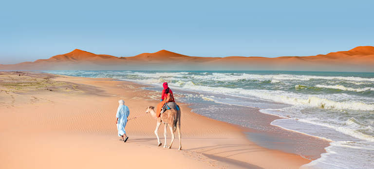 Two people in colorful clothing walking along the sandy beach, with towering orange mountains in the background and a calm, blue ocean.