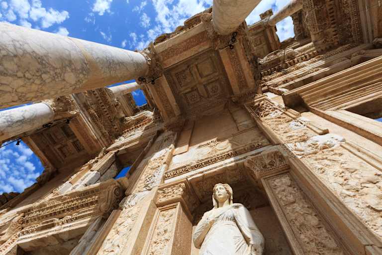 A grand, ornate stone structure with intricate carvings and a towering statue stands against a backdrop of a bright blue sky with fluffy white clouds.