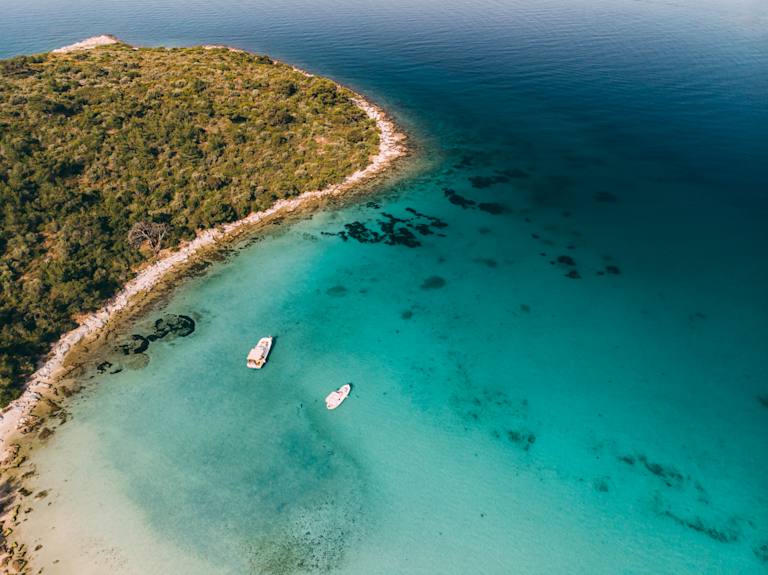 A lush, forested peninsula juts out into a stunning turquoise sea, with a few small boats anchored in the crystal-clear waters.