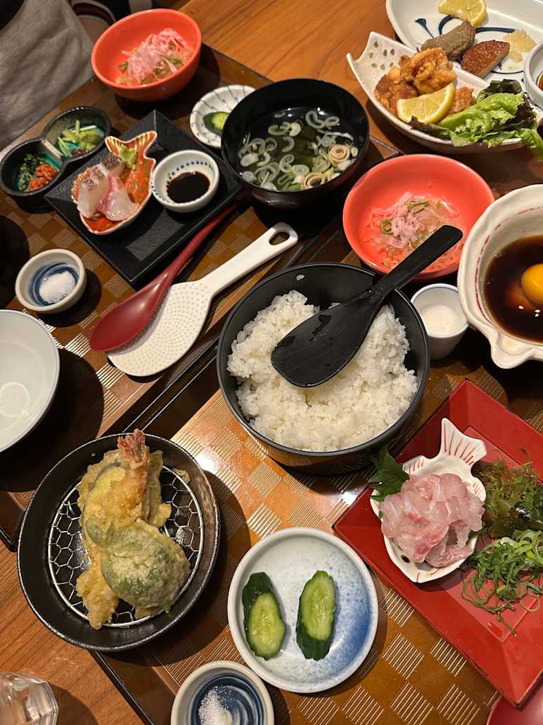 A spread of Japanese dishes, including tempura and sashimi, is laid out on a checkered tablecloth, with a bowl of rice and various small plates in the foreground and a larger platter of fried food in the background.