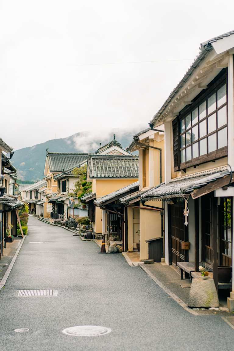 A quiet, narrow street lined with traditional Japanese houses leads towards misty mountains in the background.