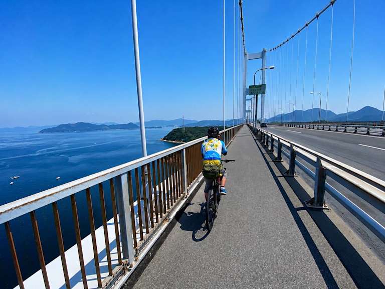 A cyclist rides on a wide bridge with a railing overlooking a calm blue sea dotted with islands under a clear sky.