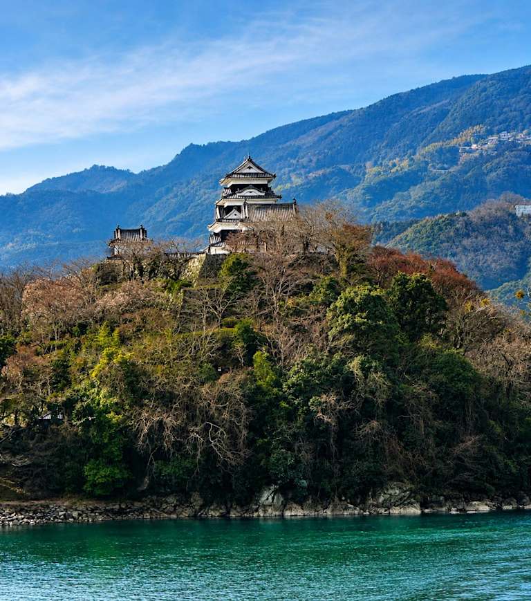 A Japanese castle stands atop a tree-covered hill in the foreground, with lush, blue mountains rising in the background under a clear sky.