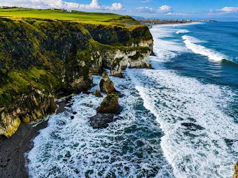 Rugged green cliffs plunge into a churning blue ocean with white-capped waves crashing against sea stacks in the foreground, while a distant coastline stretches under a bright sky.