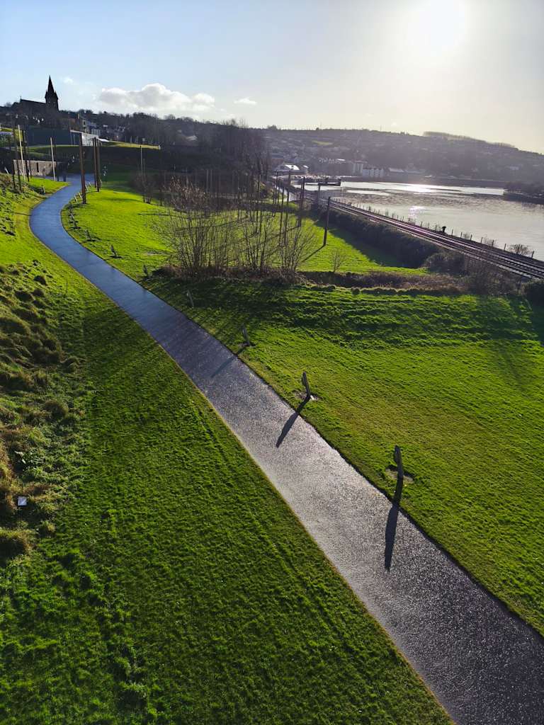 A paved path winds through a grassy hillside in the foreground, leading towards a river and distant town under a bright, hazy sun in the background.