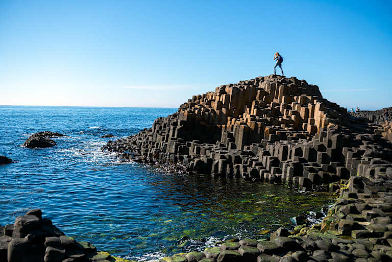 A lone figure stands atop hexagonal basalt columns in the foreground, with the blue ocean and clear sky stretching out in the background.