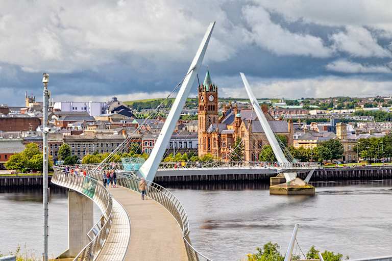 A modern pedestrian bridge curves across a river in the foreground, leading to a city skyline with a prominent clock tower in the background under a cloudy sky.