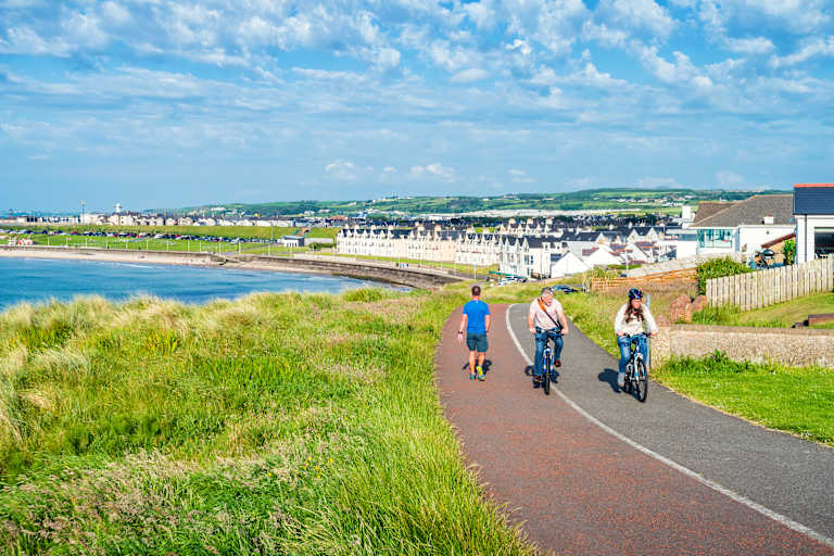 A paved path winds through grassy dunes with people cycling and walking, leading to a coastal town with white buildings under a blue sky.
