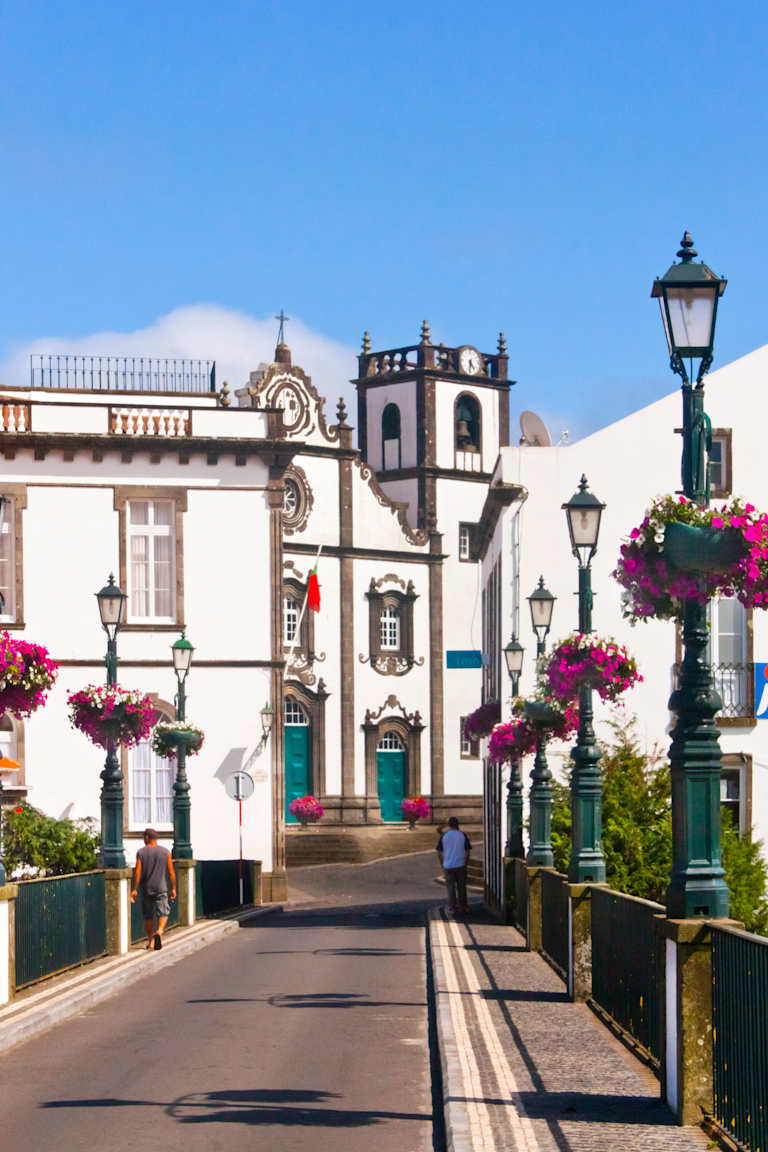 A bridge with lampposts adorned with pink flowers leads to a white building with a bell tower under a clear blue sky.