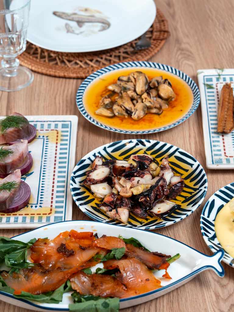 A wooden table is laden with various seafood dishes, including smoked salmon, octopus, and mussels, with a glass and a decorative plate in the background.