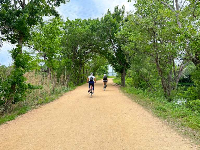Two cyclists ride down a sandy path lined with lush green trees, with a body of water visible to the right in the background.