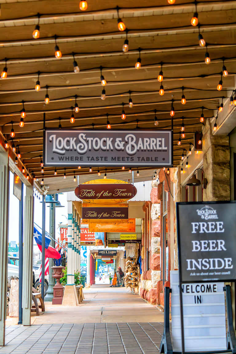 A charming storefront with a "Lock Stock & Barrel" sign and string lights overhead leads to a street lined with shops and flags.