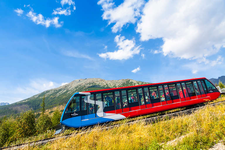 A red and blue funicular train ascends a grassy slope with a mountain range and a bright blue sky with clouds in the background.