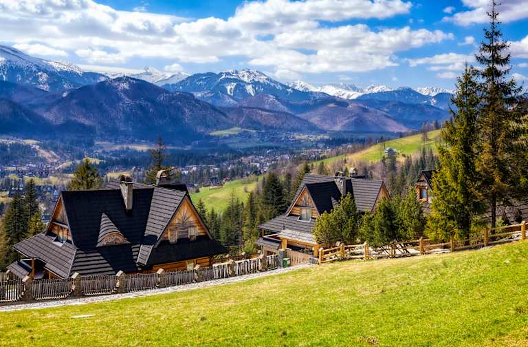 Two wooden houses with dark roofs sit on a grassy hill in the foreground, with a valley town and snow-capped mountains in the background.