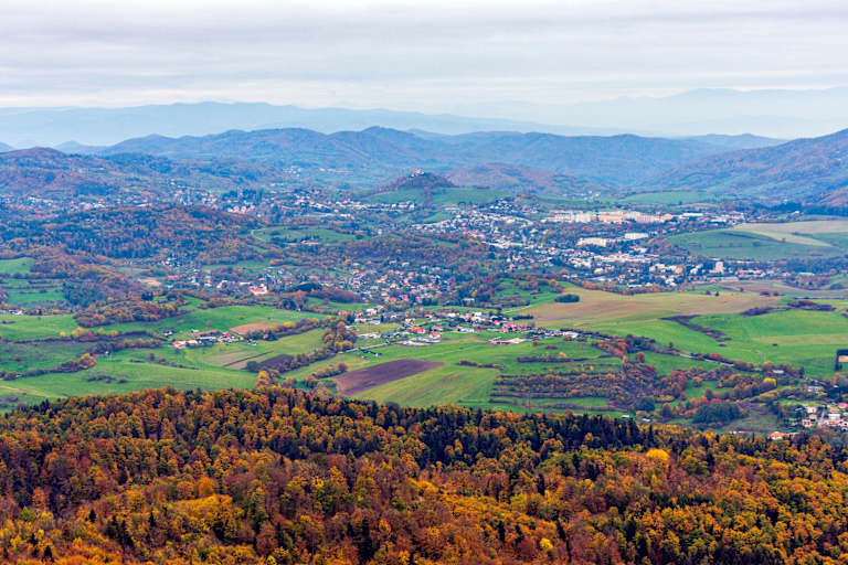 A vibrant autumn forest in the foreground gives way to a sprawling town nestled among rolling green hills and distant blue mountains.