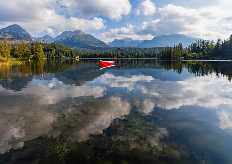 A bright red boat floats on a calm lake reflecting the cloudy sky and distant mountains.