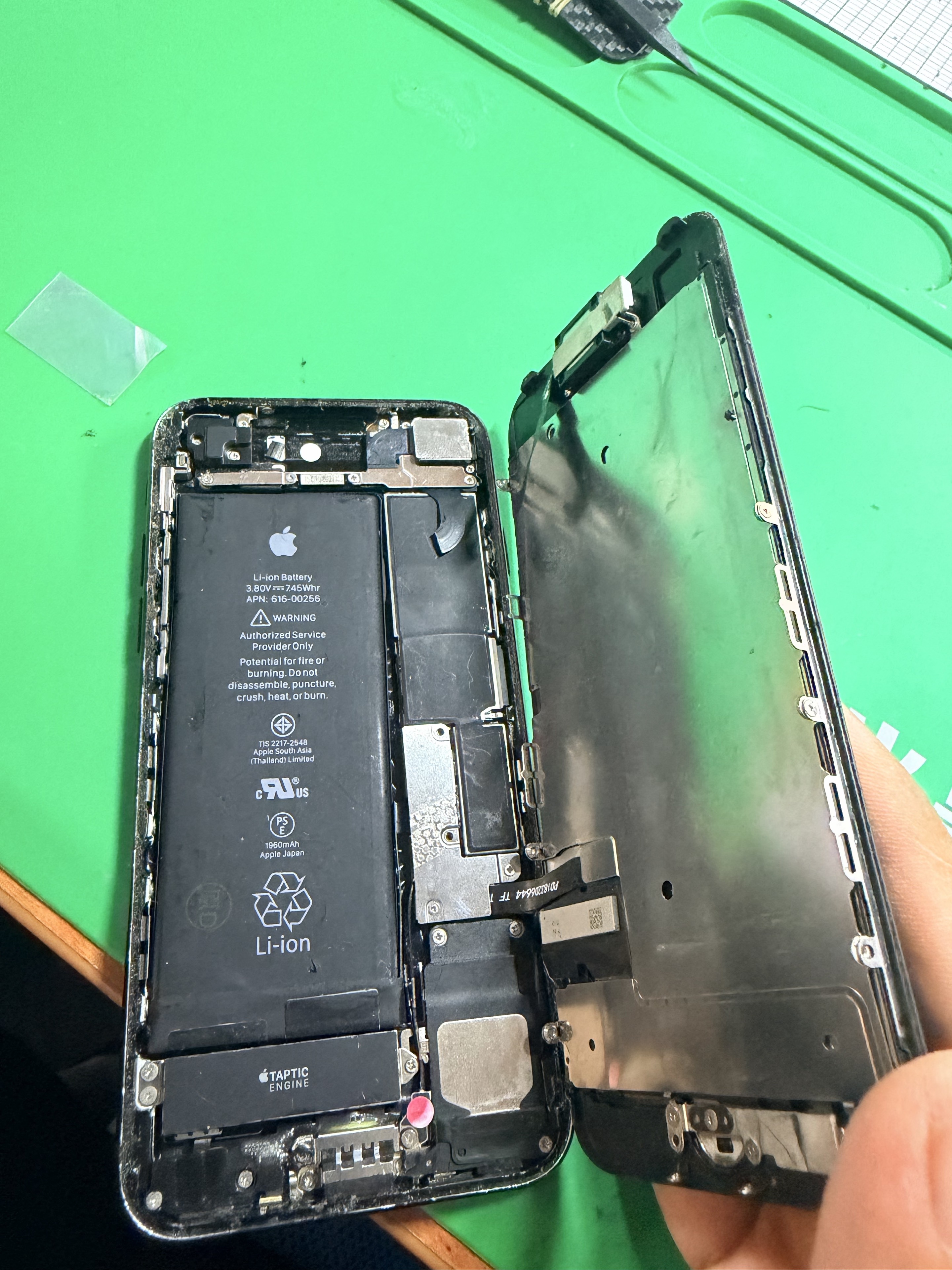 iPhone dengan skrin dibuka dan bateri dalaman kelihatan di atas alas kerja hijau