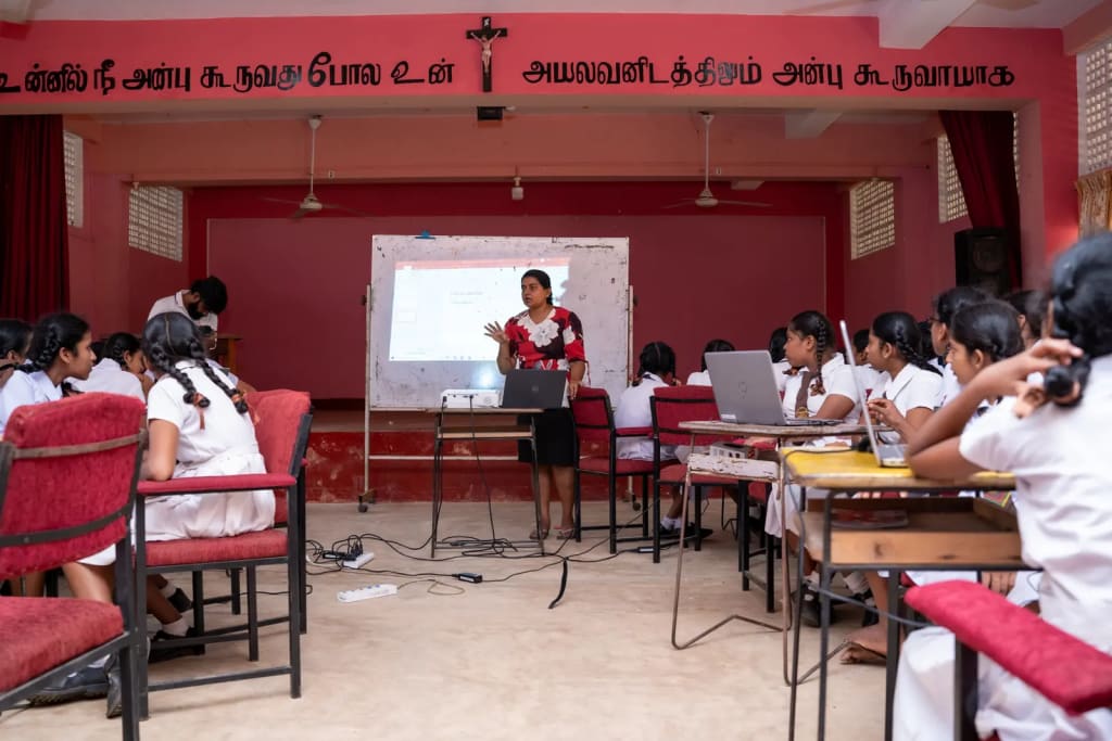 Students work in groups at laptops while a facilitator presents at the front of a school hall.