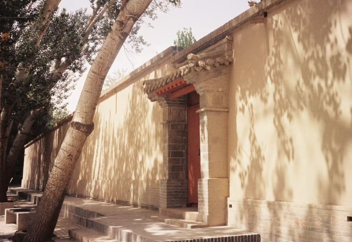 A sunlit traditional courtyard wall and red doorway beneath leafy trees