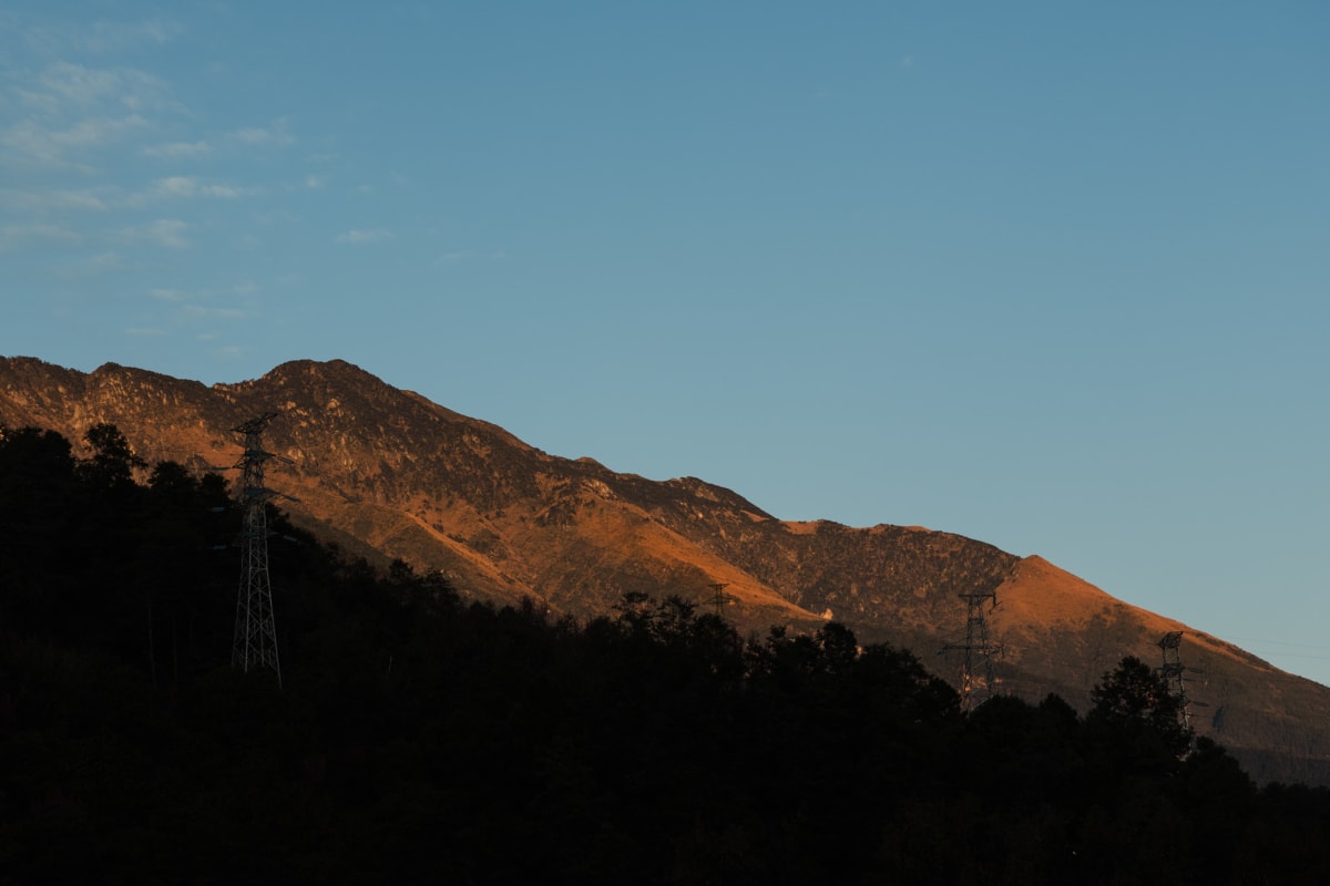 A sunlit mountain ridge rising above dark forest beneath a clear blue sky