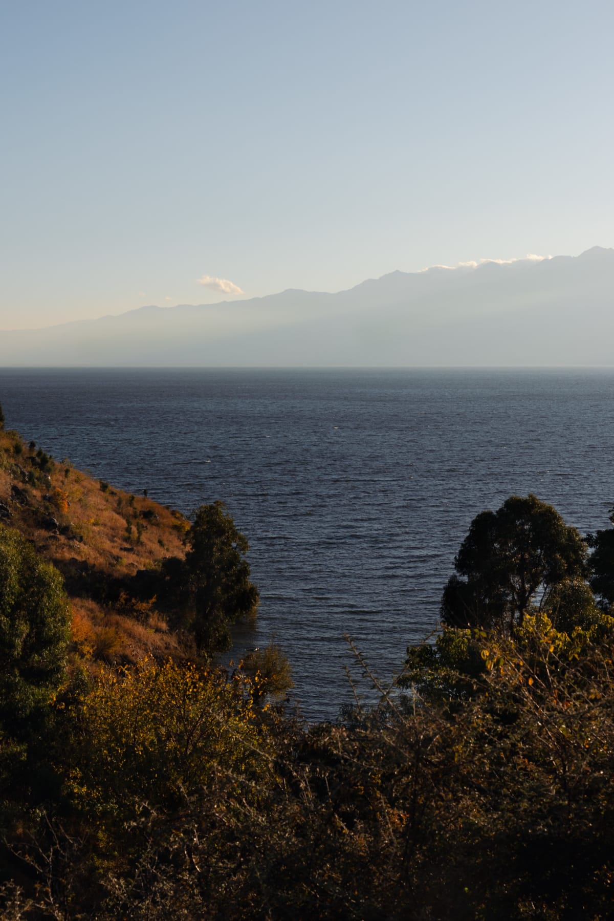 A dark blue lake framed by sunlit foliage with hazy mountains beyond