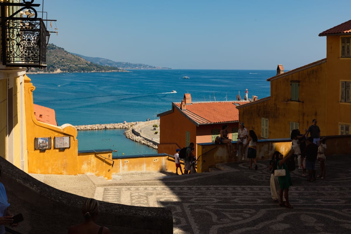 A sunlit coastal town descending toward a vivid blue Mediterranean harbour