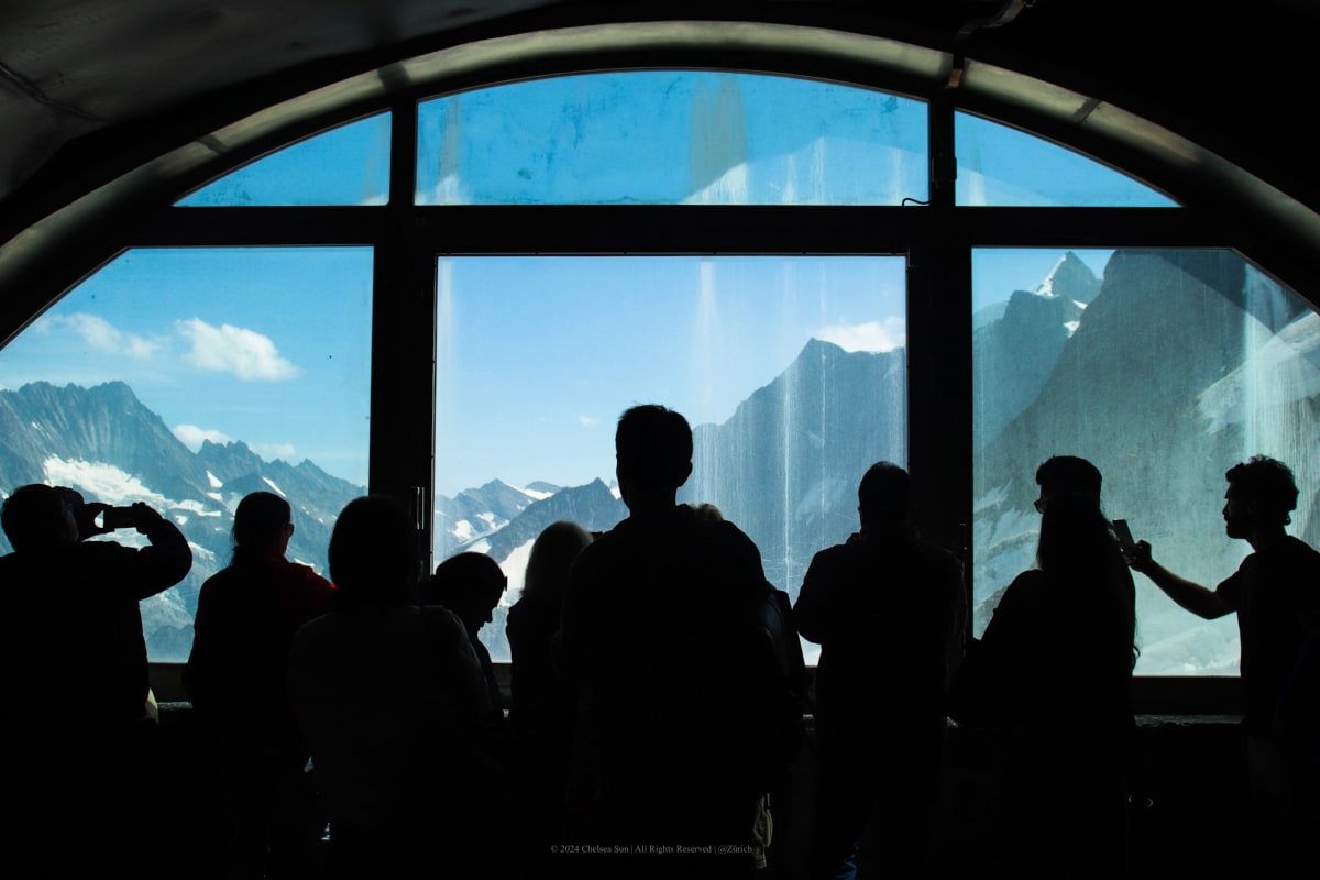Visitors silhouetted before a panoramic window overlooking alpine peaks