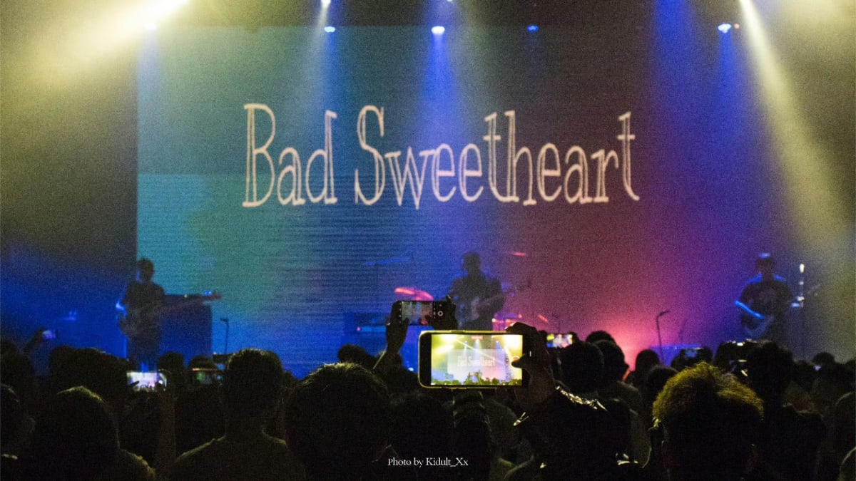 A band performing beneath blue and yellow lights as the audience records the show