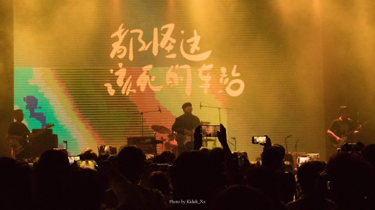 Musicians performing beneath amber lights and handwritten characters on screen