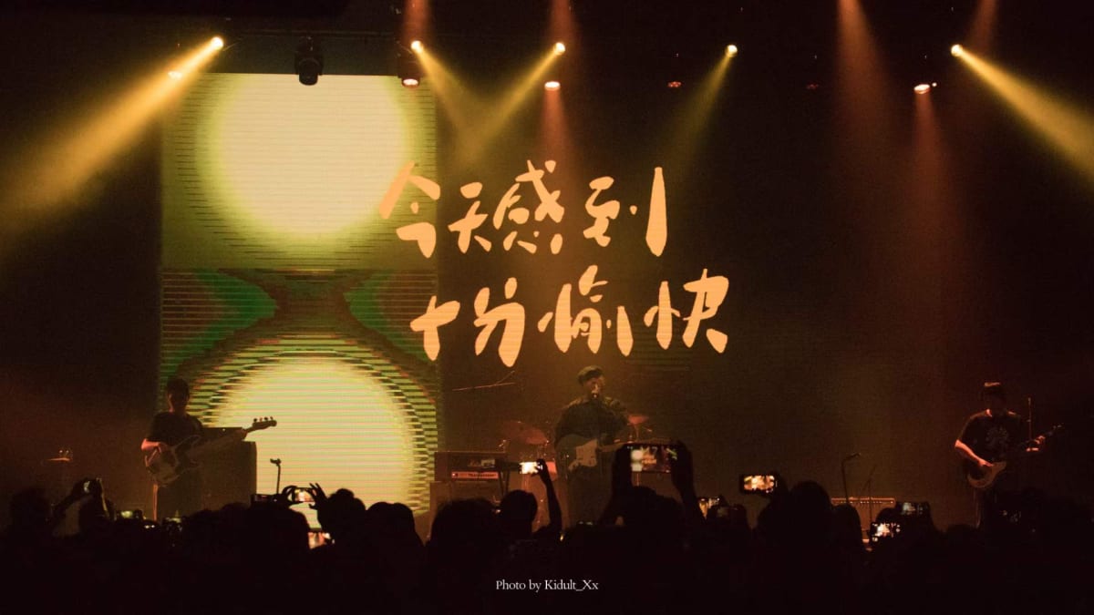 A band performing under amber spotlights before a sea of raised phones