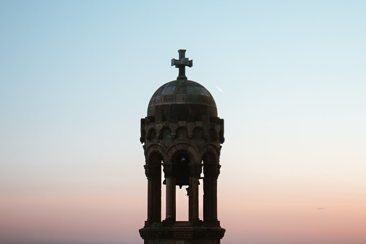 A stone bell tower topped by a cross against a pastel sky