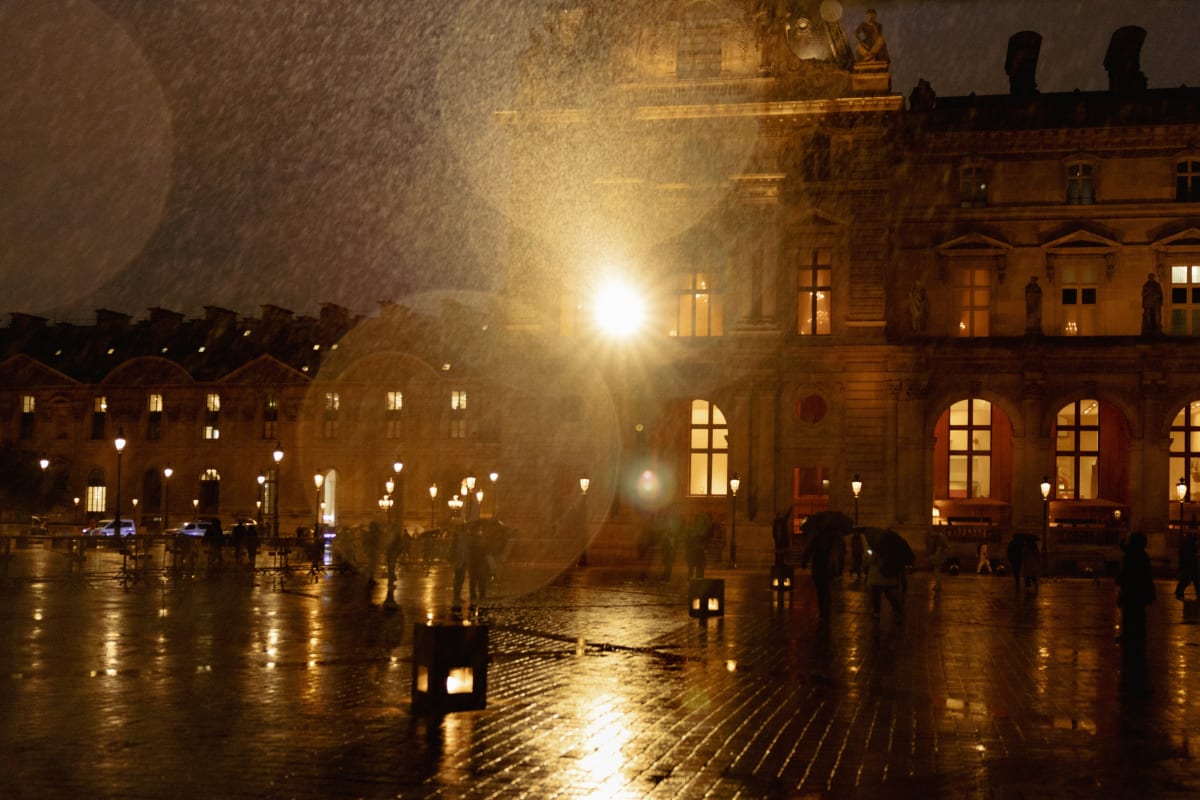 Rain falling through lamplight across a reflective Paris courtyard