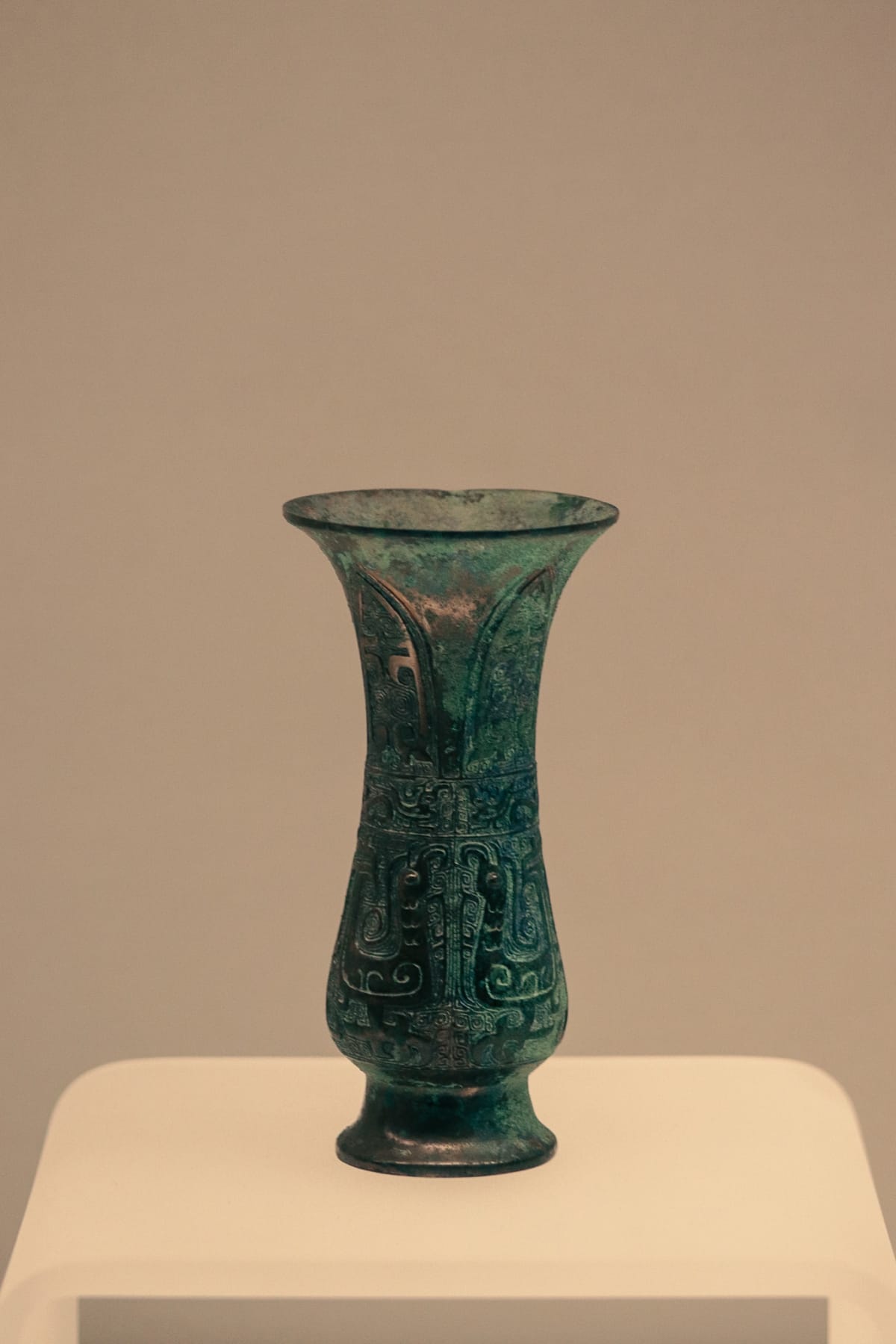 A slender green bronze vessel displayed on a pale pedestal