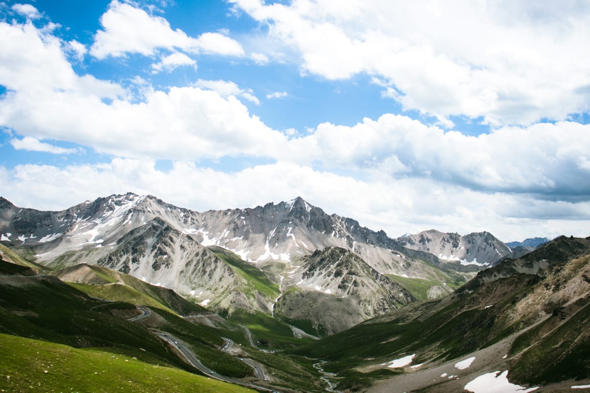 A winding mountain road through a green valley beneath snow-streaked peaks