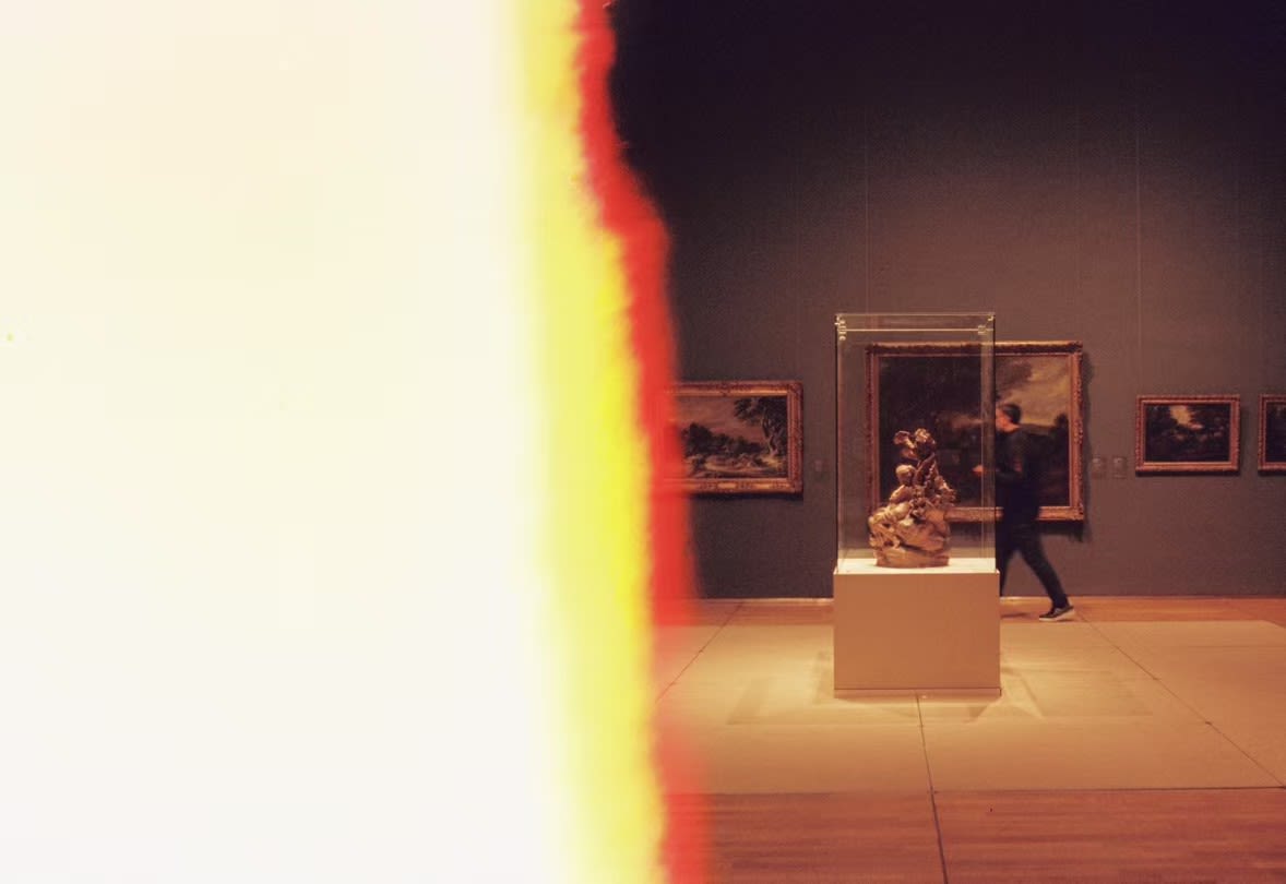 A film-burn flare revealing a sculpture and passing visitor inside a museum gallery